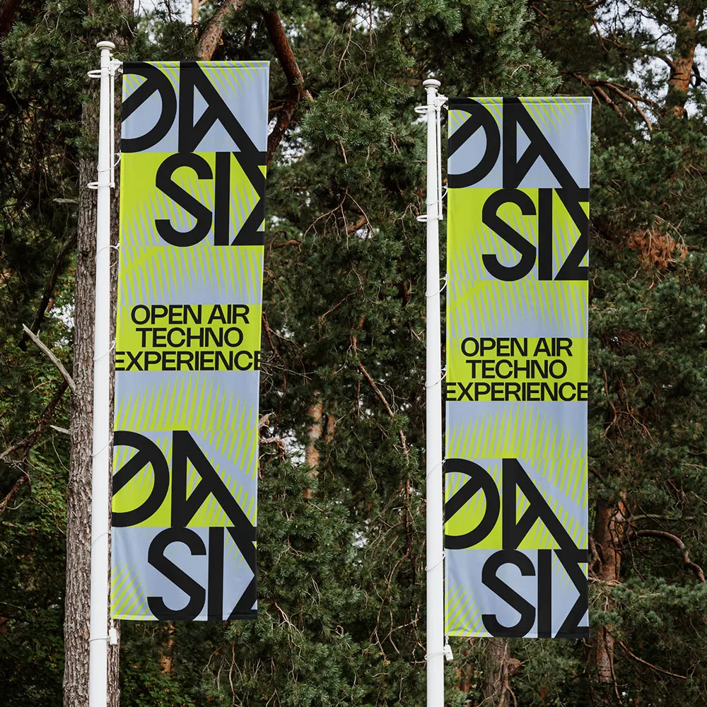 Two vertical banners with abstract green and gray design and text saying 'OPEN AIR TECHNO EXPERIENCE' hanging on white poles in front of pine trees.